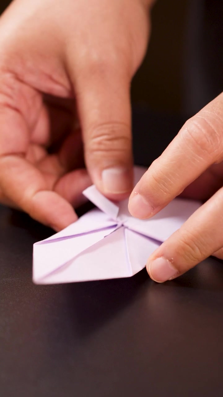 Close-up hands carefully folding purple paper into origami ship, soft lighting, steady camera angle