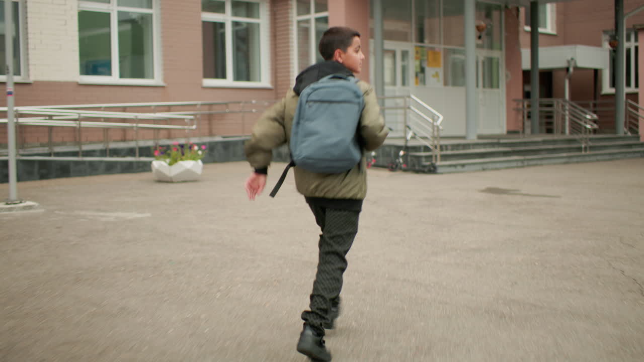 학교 입구, 코트야드 표시가 보이는 바닥, 학업 분위기와 젊음의 활기가 느껴지는 건물 배경을 향해 뒤돌아보며 가방을 메고 달리는 학생의 뒷모습