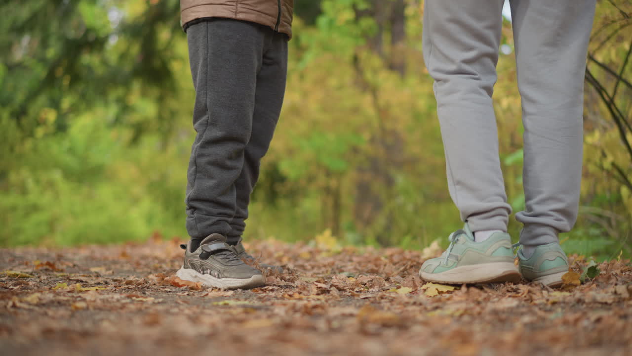 close low angle view of child and adult standing still on forest trail during walk, focusing on legs and ground, surrounded by blurred foliage in tranquil autumn landscape
