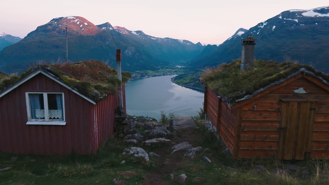 Tomada em dolly da câmera se movendo entre duas pequenas casas, revelando uma bela paisagem de lago e montanhas