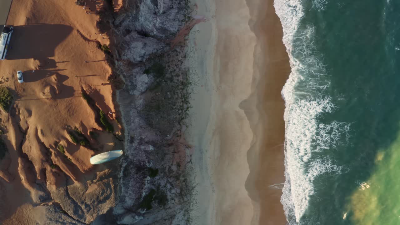 Aerial drone bird's eye top view following a paraglider flying over the Cliffs of Cacimbinhas in Pipa, Brazil Rio Grande do Norte as small waves crash into the beach on a warm summer day