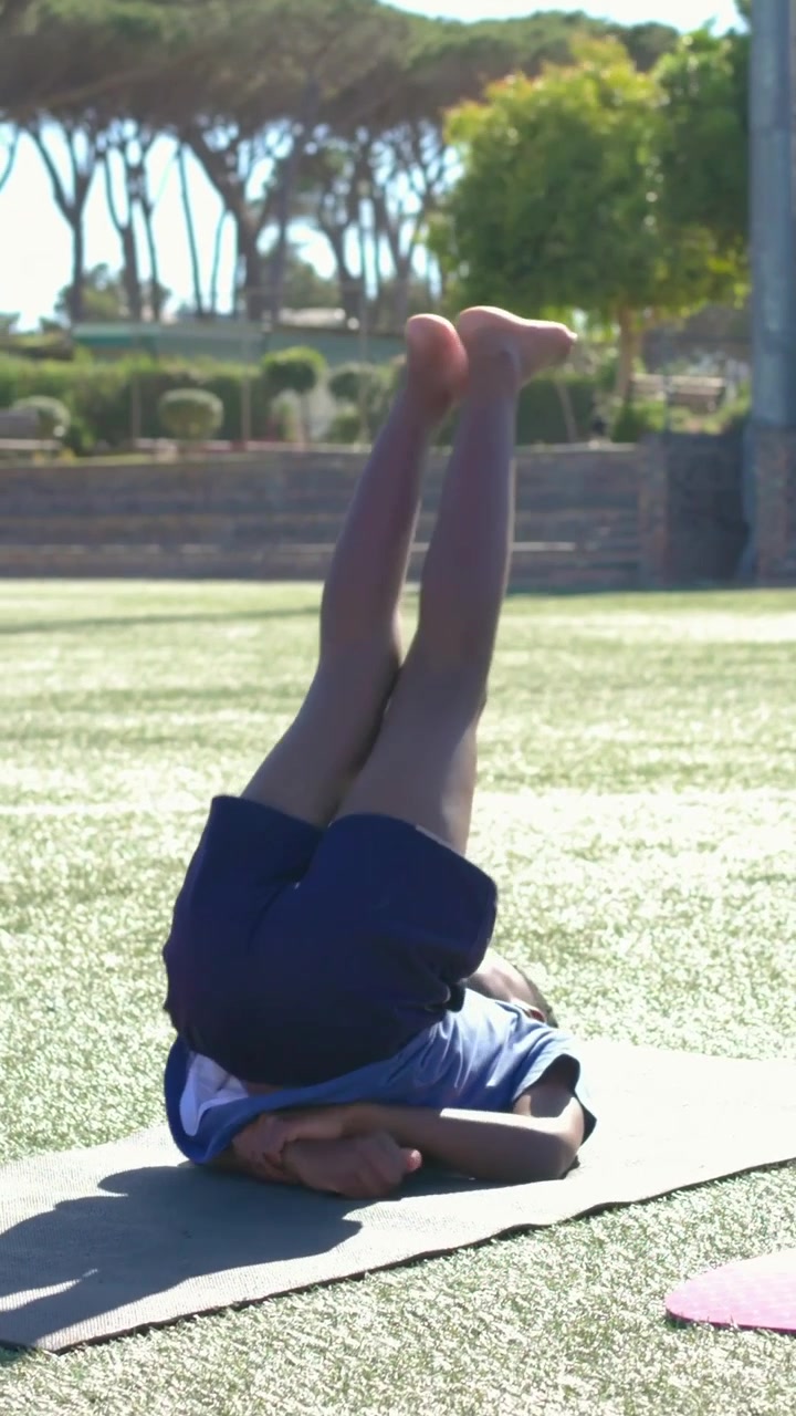 Video verticale: Un ragazzo con una maglietta blu rotola da una verticale sulle spalle e si allunga su un tappetino da ginnastica scolastico per la lezione.