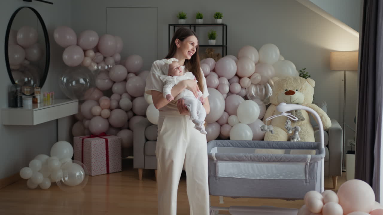 Donna in bianco culla un neonato circondata da palloncini pastello e articoli per la cameretta