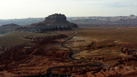 A-tilting-up-aerial-drone-desert-scene-with-a-winding-narrow-road-surrounded-by-red-rocks-and-sand-with-large-red-and-white-butte-rock-formations-in-the-background-in-Goblin-Valley-Utah-State-Park