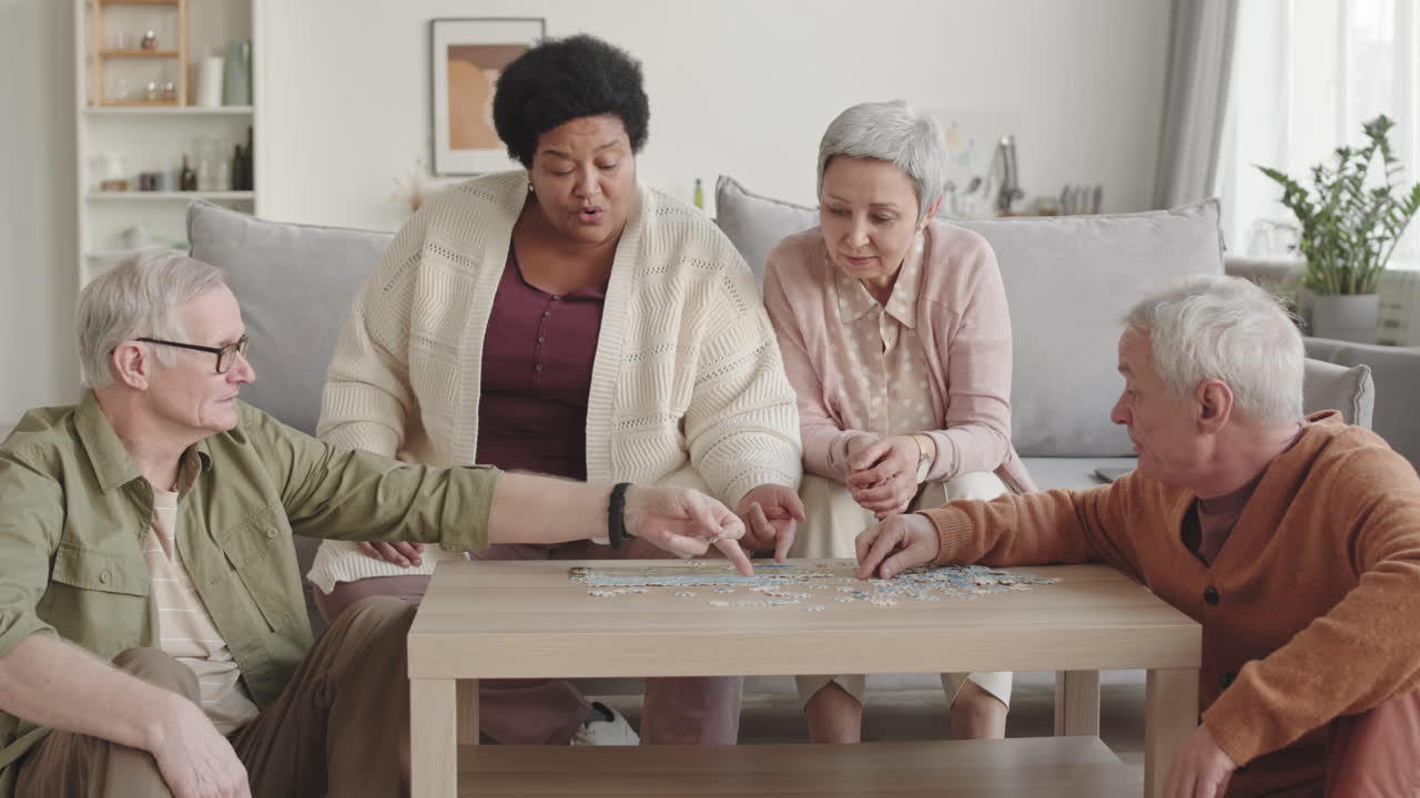 Medium long of Asian and African women and two senior Caucasian man sitting in living room at coffee table, smiling, chatting, doing puzzle