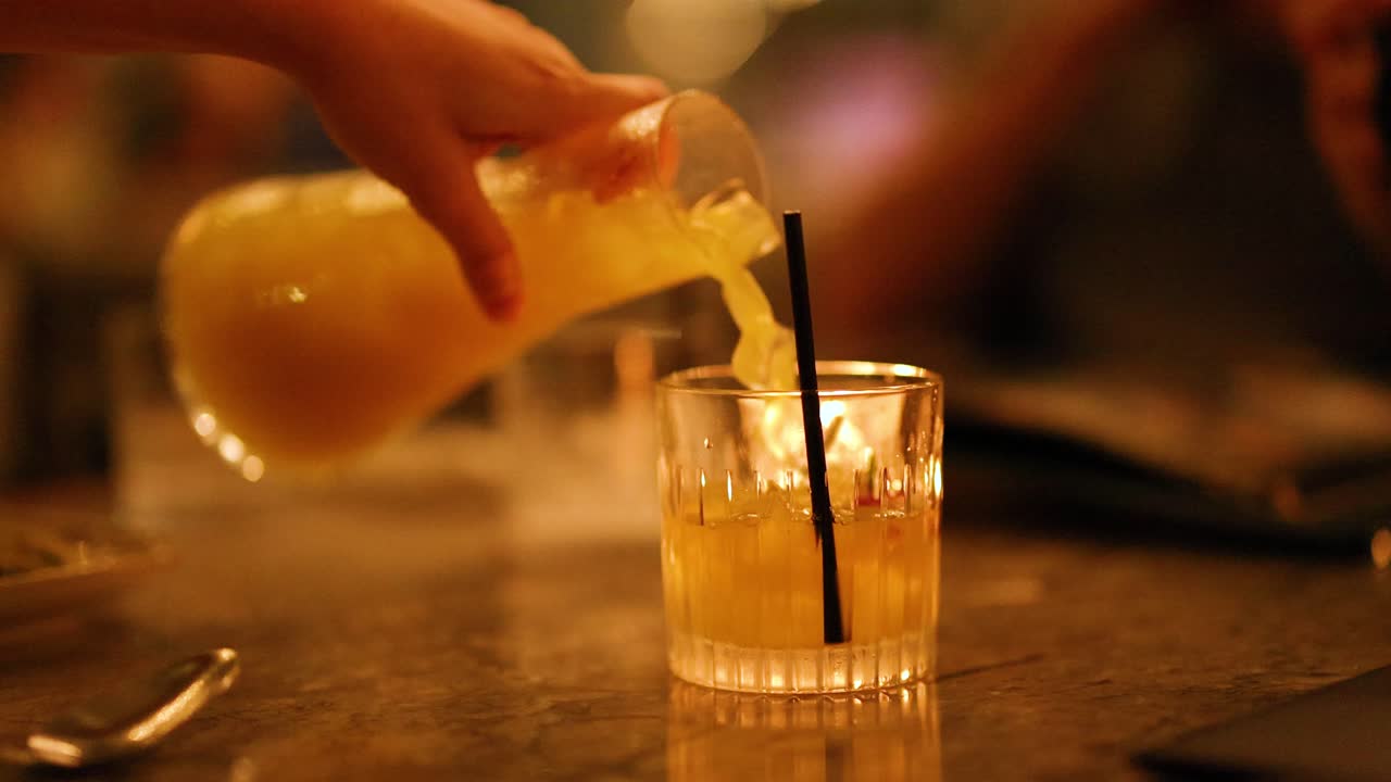 A hand pours a vibrant cocktail into a glass in a softly lit bar setting, creating a warm, inviting atmosphere