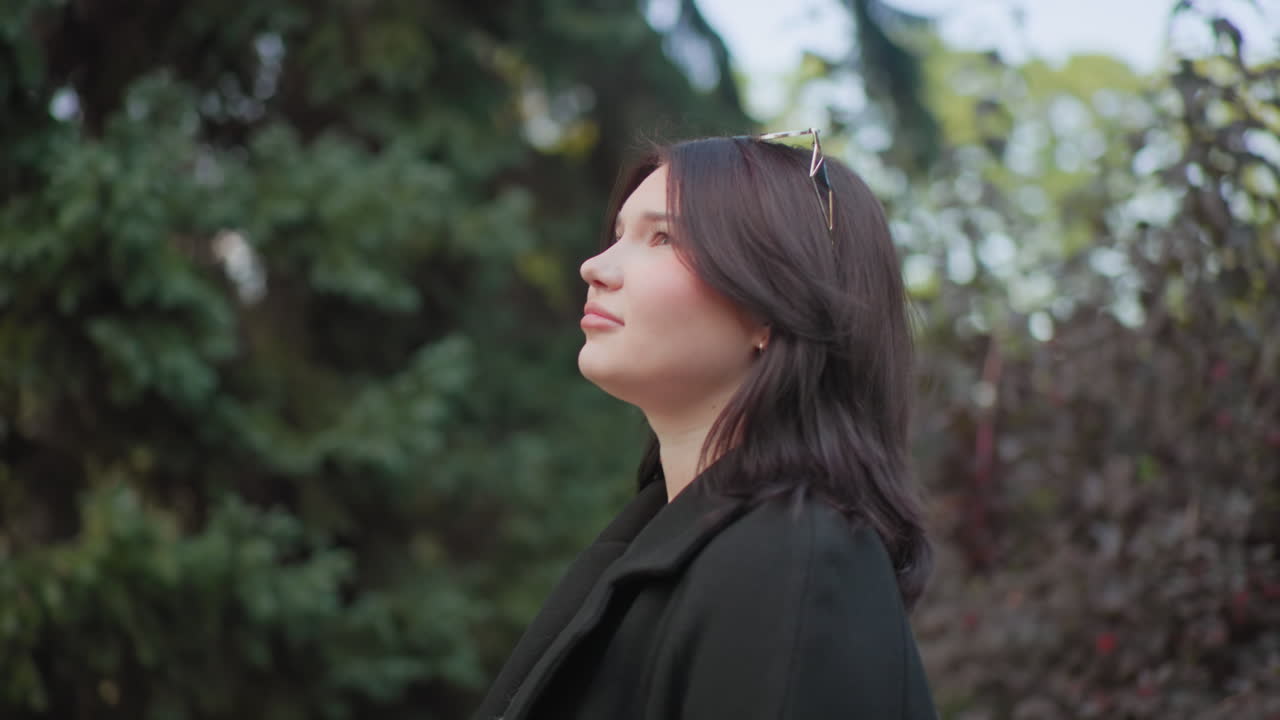 가을을 사랑하는 사람이 공원 산책로를 거닐고, 머리에는 선글라스를 끼고, 어두운 코트와 스웨터를 입고, 부드러운 녹색 아래에서 천천히 걷는 모습의 측면 뷰, 자연 속에서 평온한 분위기, 나무에 둘러싸인 야외 프로필, 보케 빛