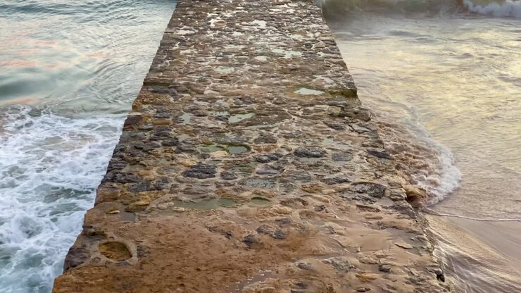 Primer plano de la entrada al pontón oceánico con un enorme atardecer de fondo en la playa de Cascais con algunas olas rompiendo.