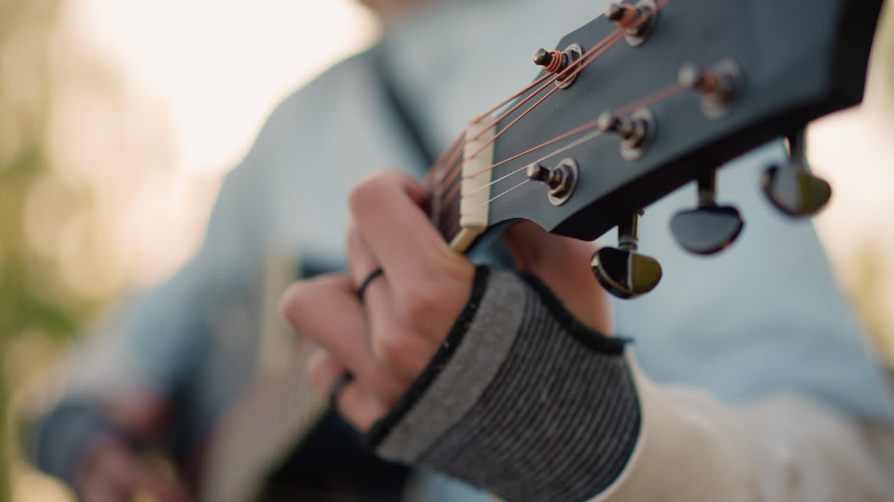 white tutor fingerpicking guitar headstock closeup, ringed fingers tuning strings, knit glove sleeve visible, focused technique, shallow depth with bokeh, tutorial atmosphere, precise hand movements