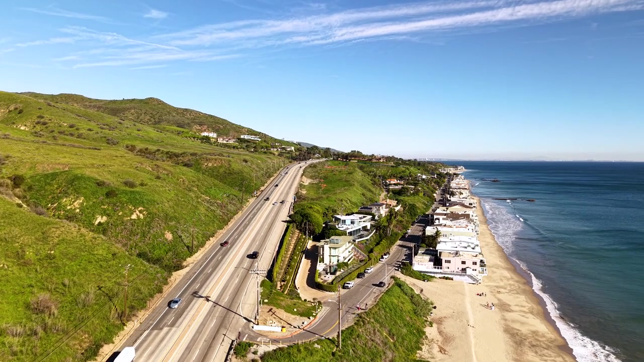Liikenne Pacific Coast Highwaylla virtaa ohi Malibu Beachin, kun drone seuraa autojen rinnalla ajaen ikonista Kalifornian rannikkoreittiä valtameren ja vihreiden kukkuloiden välissä. Corral Beachin näkymä.