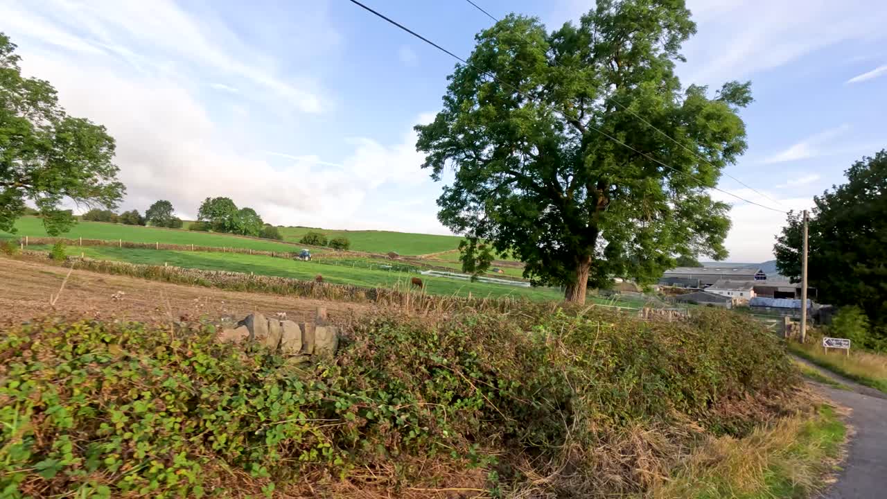 A vehicle travels along a narrow country road bordered by fields, stone walls, and trees in the English countryside under soft evening sunlight