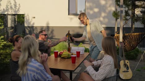 Happy-blond-guy-brings-a-package-of-pizza-and-bottles-of-drinks-to-the-table-during-a-shared-lunch-in-the-courtyard-of-a-country-house