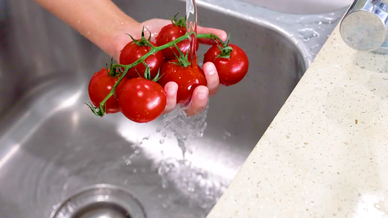 Hands rinse tomatoes under running water in a kitchen sink, highlighting cleanliness and freshness with bright lighting