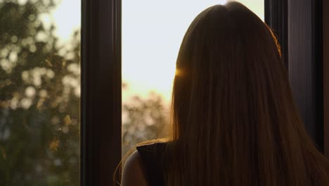 toma mediana de mano de una mujer mirando por la ventana la puesta de sol y los árboles antes de volverse para mirar a la cámara mientras sostiene un vaso de whisky