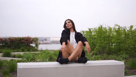 gorgeous brunette model sitting down on grey bench in london city park near to a river relaxing posing wearing fashionable trendy black jacket and trainers looking cute moving shot cloudy weather