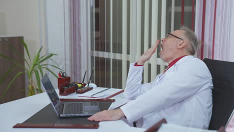 tired doctor in front of laptop. senior male specialist in medical coat sitting in hospital office and having online consultation with a patient. remote medicine.