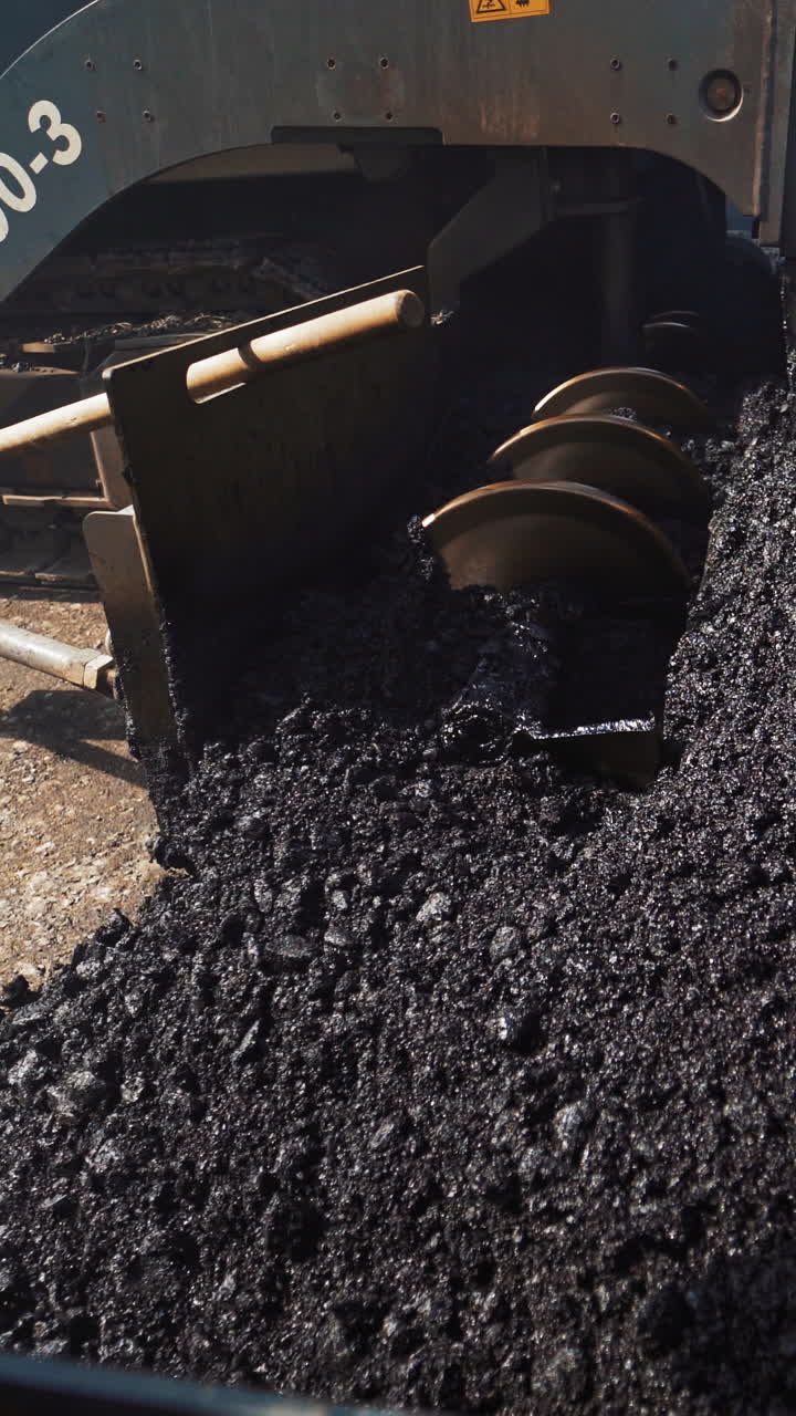 Metal construction of a heavy paver machine pouring out hot asphalt on the road. Asphalt spreader at work. Large truck laying black bitumen. Close-up. Vertical video