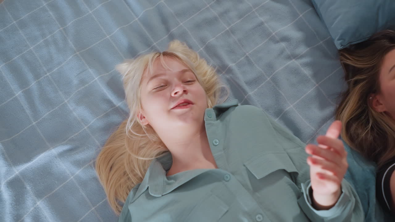 Blonde girl in mint pajama laughs and sings with eyes closed while lying on bed beside smiling friend, both express joy with raised hands, creating fun, carefree atmosphere in cozy bedroom setting