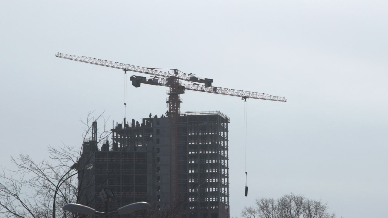 Tower Crane Over Highrise Construction Site, Gray Sky And Bare Trees, Crane Hoisting Concrete Slab . Media