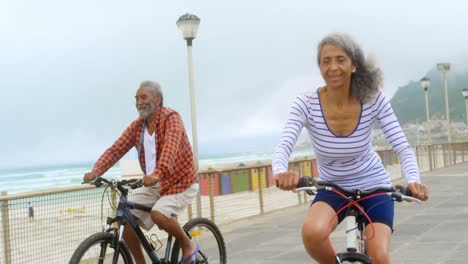 Vista-Frontal-De-Una-Pareja-Afroamericana-Senior-Activa-Montando-Una-Bicicleta-En-El-Paseo-Marítimo-4k