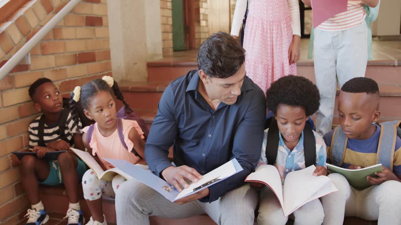 Diverse male teacher and children sitting on stairs reading books in elementary school, slow motion