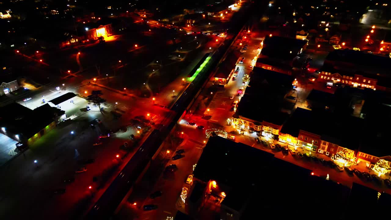 An aerial drone shot captures downtown Hinsdale, Illinois at night, adorned with festive holiday lights, showcasing illuminated buildings, busy streets, and railway tracks