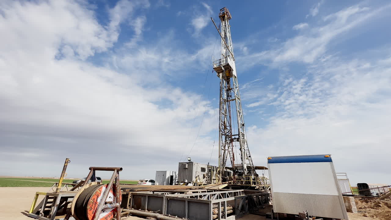 Metal details and pipes at the site for natural resources production. High tower for frilling gas at backdrop.