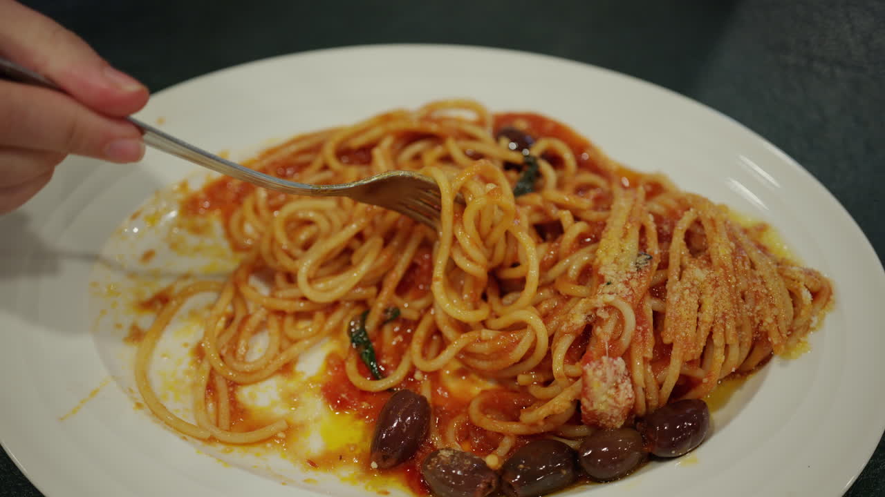 Potret dekat seseorang yang menggulung spaghetti alla puttanesca dengan garpu, memperlihatkan saus tomat kaya rasa, buah zaitun, dan keju parut.