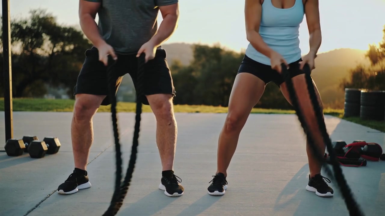 Et træningspar i shorts griber fat i battle-ropes og laver skiftevis bølger på en betonplade med vægtskiver.