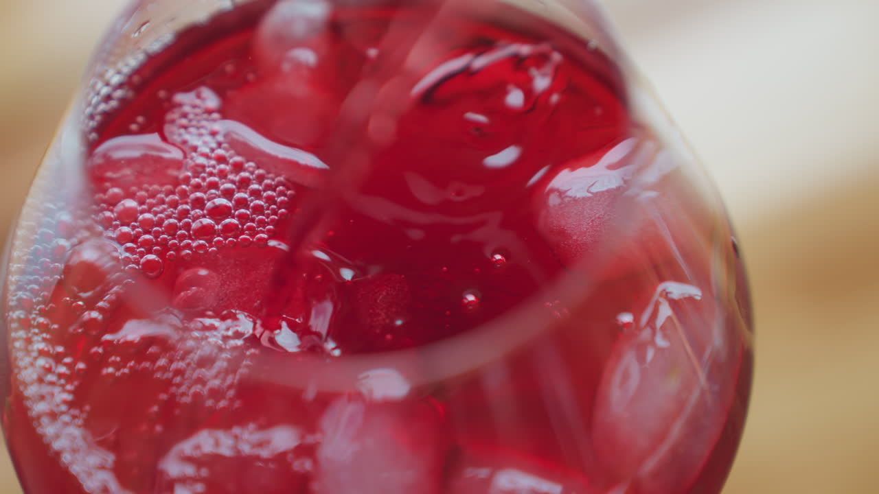 Close up of vibrant red wine pouring into transparent glass filled with ice cubes, creating bubbling effect and rich texture as chilled drink swirls around frozen pieces in refreshing motion