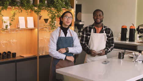 Two-Multiethnic-Waiters-With-Crossed-Arms-Smiling-At-Camera-While-Standing-Behind-Counter-In-A-Coffee-Shop