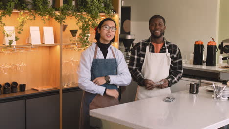 dos camareros multiétnicos con los brazos cruzados sonriendo a la cámara mientras están detrás del mostrador en una cafetería