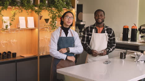 two multiethnic waiters with crossed arms smiling at camera while standing behind counter in a coffee shop