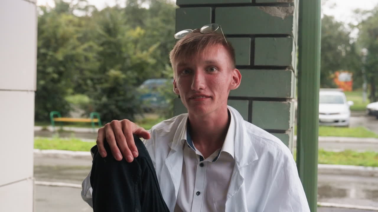 Portrait view of young man in white shirt and coat sitting with relaxed posture, smiling warmly outdoors near wall, with glasses resting on head, reflecting casual human expression and natural moment