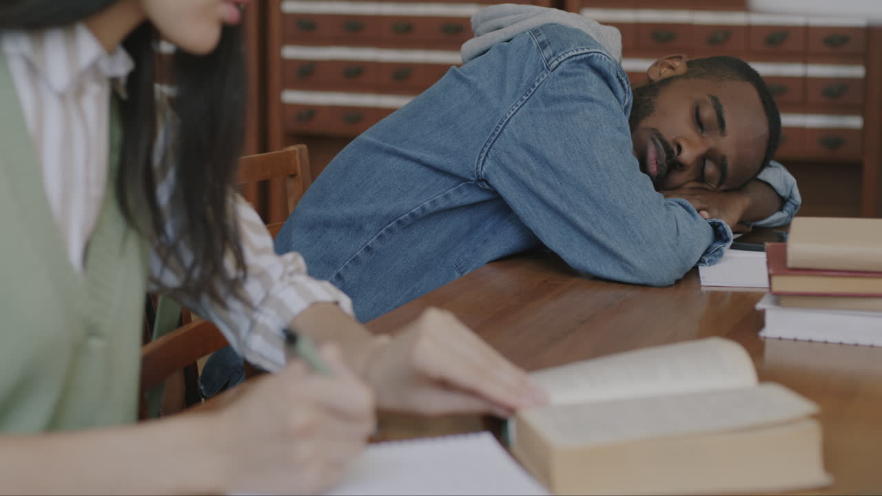 Student Sleeping in Library While Another Studies