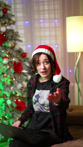 cheerful caucasian woman in santa cap looks at her laptop. happy lady waves to camera calling up. christmas tree at backdrop. vertical video.