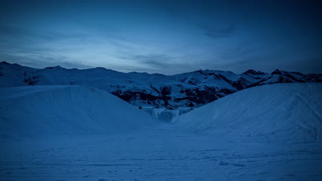 sunset time lapse from within half pipe at ski resort - snowy mountains and snow groomers preparing slopes with lights on