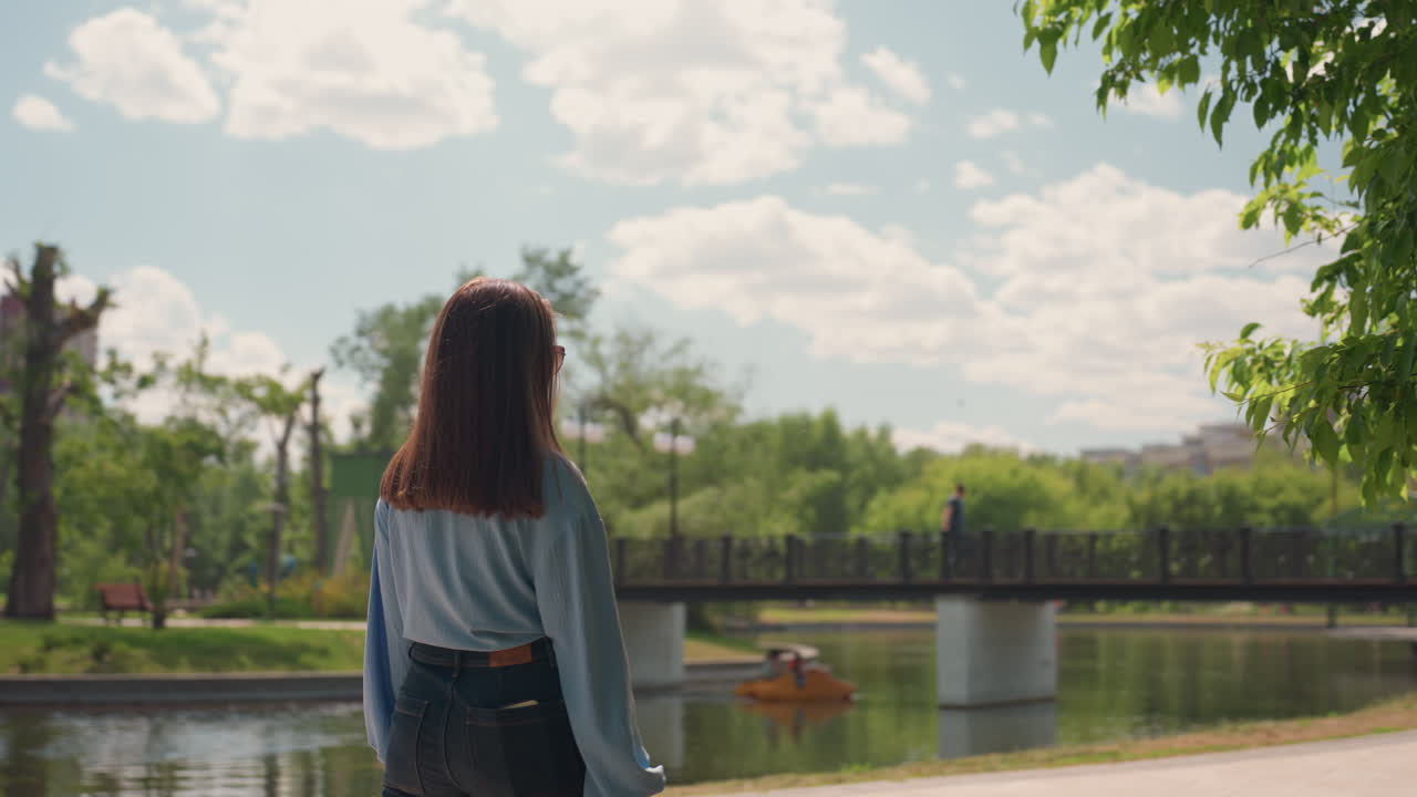 잔잔한 물가 근처 데님을 입은 젊은 여성, 다리 옆 잔잔한 호숫가에 서 있는 백인 여성, 흐린 햇빛 아래 평화로운 호숫가 풍경을 즐기는 데님 차림의 젊은 여성