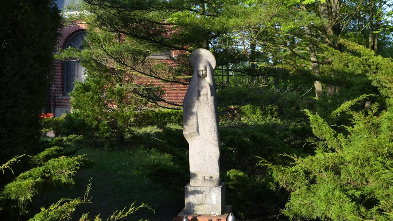A stone statue of the Virgin Mary standing at the Central Cemetery in Szczecin surrounded by lush greenery and trees, in front of a red brick chapel. Authentic movement, no stabilizer