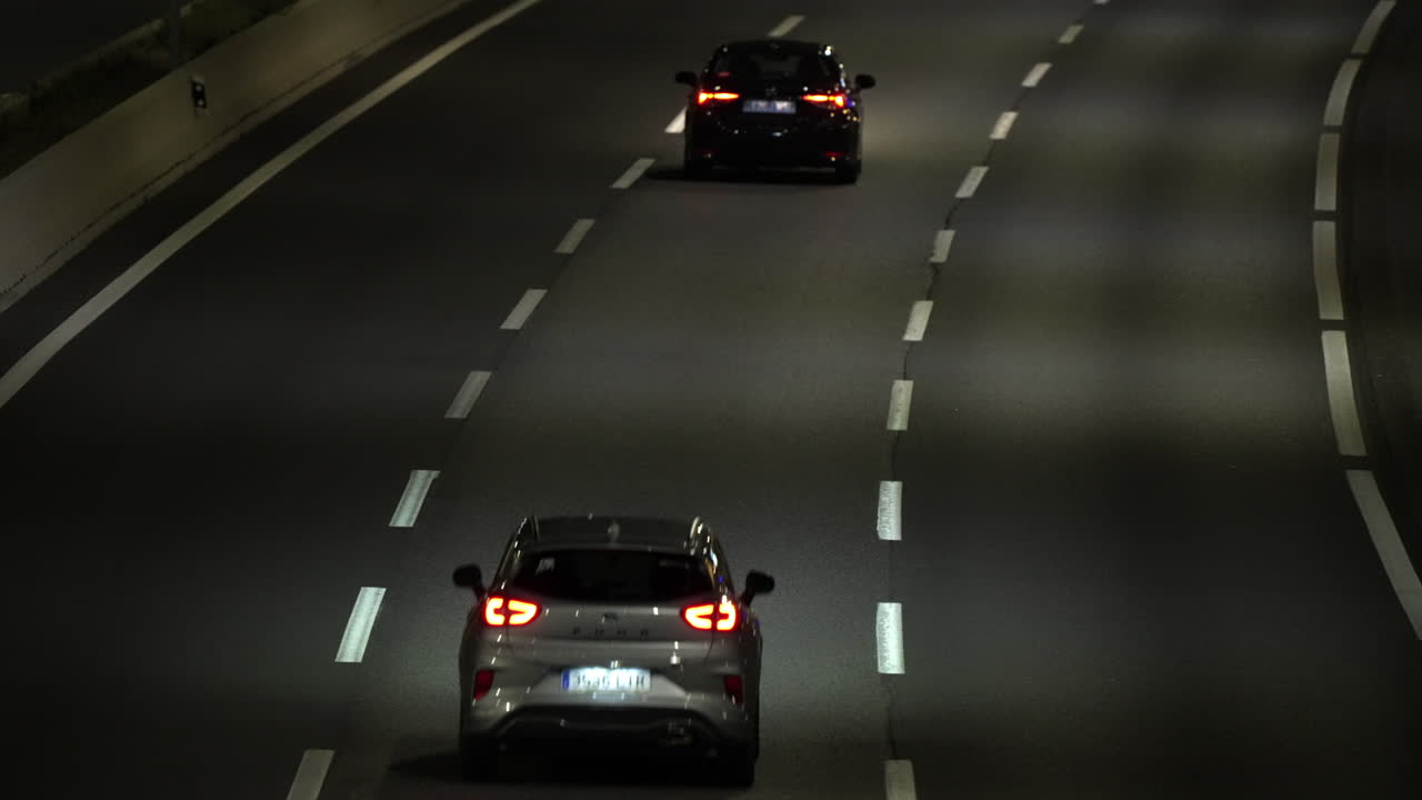 Cars are driving on a highway at night, their headlights illuminating the road ahead and taillights visible from behind as they travel in the same direction