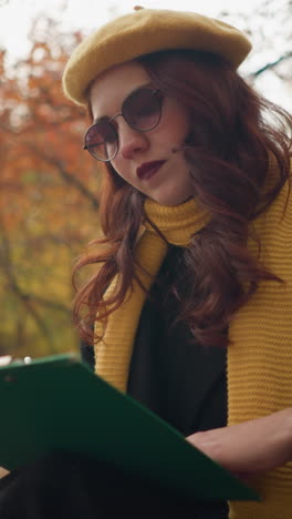 dama elegante sentada centrándose en su libro con una cubierta verde, ajustando su cabello, rodeada de hojas secas de otoño y la luz del sol brillando a través de los árboles