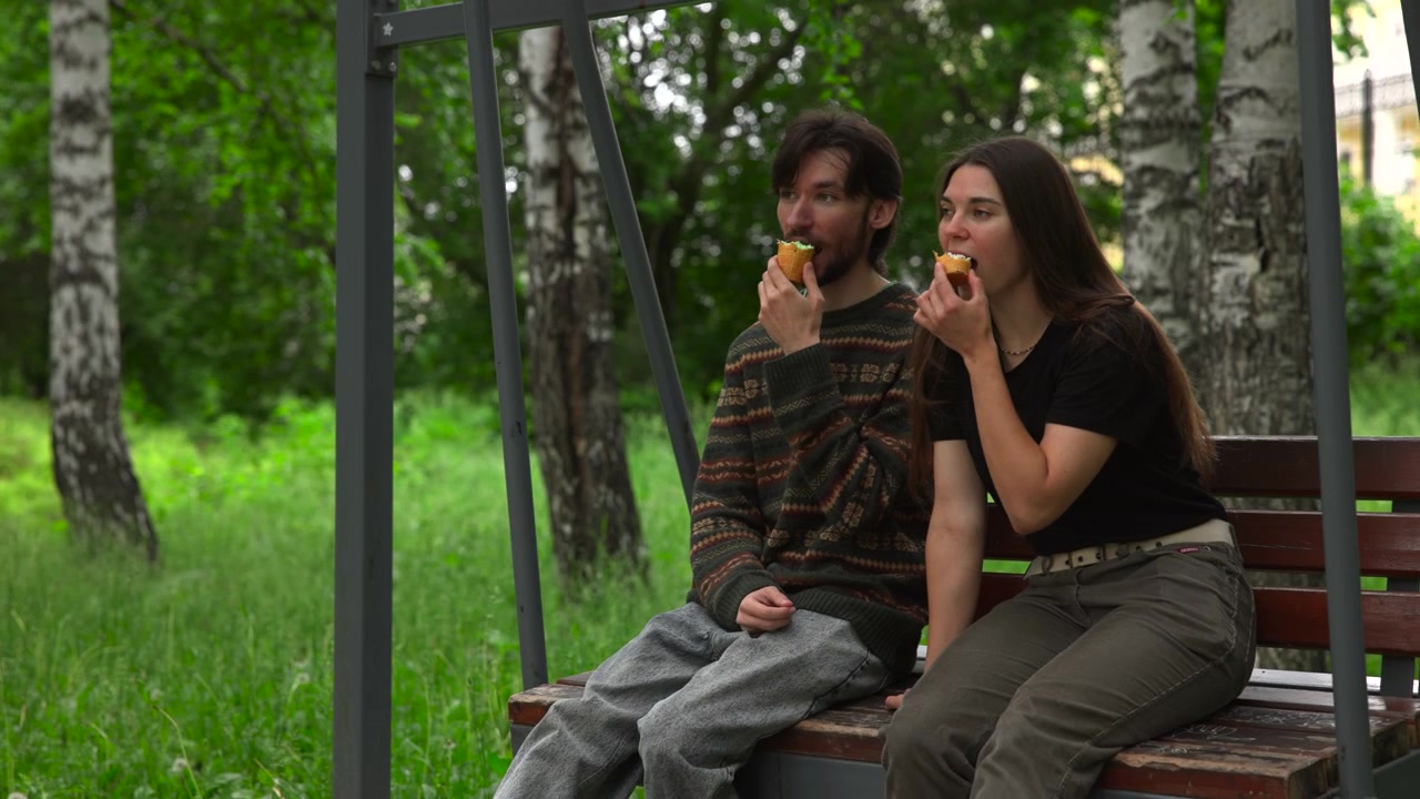 Dua orang muda duduk berdampingan di bangku ayun kayu di bawah naungan logam di hutan pohon birch berumput, memegang kue kering isi krim, mengunyah, bersantai, dan menikmati istirahat tenang di luar ruangan