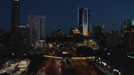 Aerial-drone-shot-slowly-descending-during-sunset-blue-hour-looking-out-on-the-skyscrapers-of-Buckhead-in-Atlanta,-Georgia-while-cars-and-a-subway-pass-by-on-the-highway-below