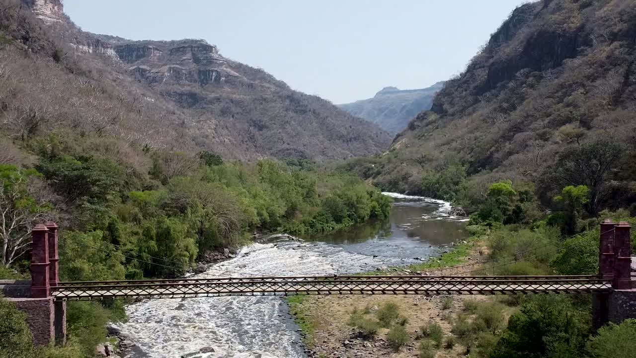 drone aéreo volando lejos de un puente colgante que cruza un río en un cañón