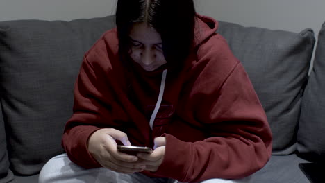 a teenage youth dressed in a hoody sitting on a sofa at home, the young girl focused on her mobile phone as she messages with a group of friends