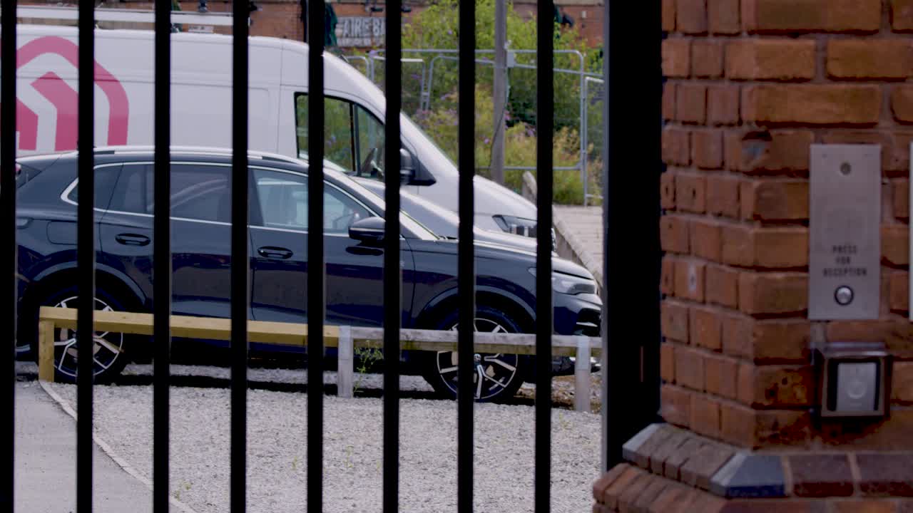 En sort metalsikkerhedsport åbner for at afsløre parkerede køretøjer, herunder en varevogn og SUV, på en grusplads ved siden af en murstenssøjle i dagslys. Statisk kamera, naturlig belysning.