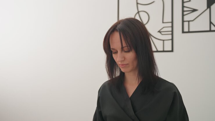 Donna caucasica in accappatoio nero, tranquilla scena mattutina di cura della persona, che sistema i capelli con movimenti delicati, parete neutra dello studio con line art minimalista, luce naturale tenue, atmosfera contemplativa, trucco discreto, pelle