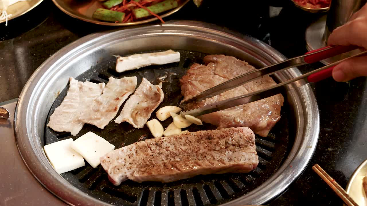 Hand uses tongs to grill garlic and pork belly slices on tabletop Korean barbecue grill