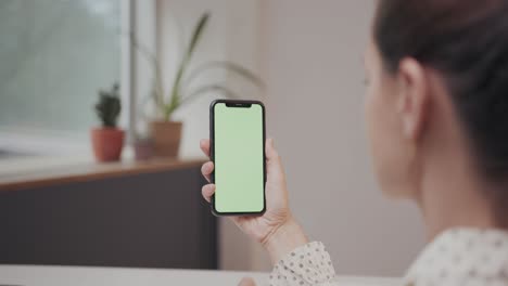 Mujer-De-Negocios-Caucásica-Deslizando-El-Dedo-Por-Un-Teléfono-Inteligente-De-Pantalla-Verde,-Mientras-Está-Sentado-En-Un-Escritorio-De-Oficina