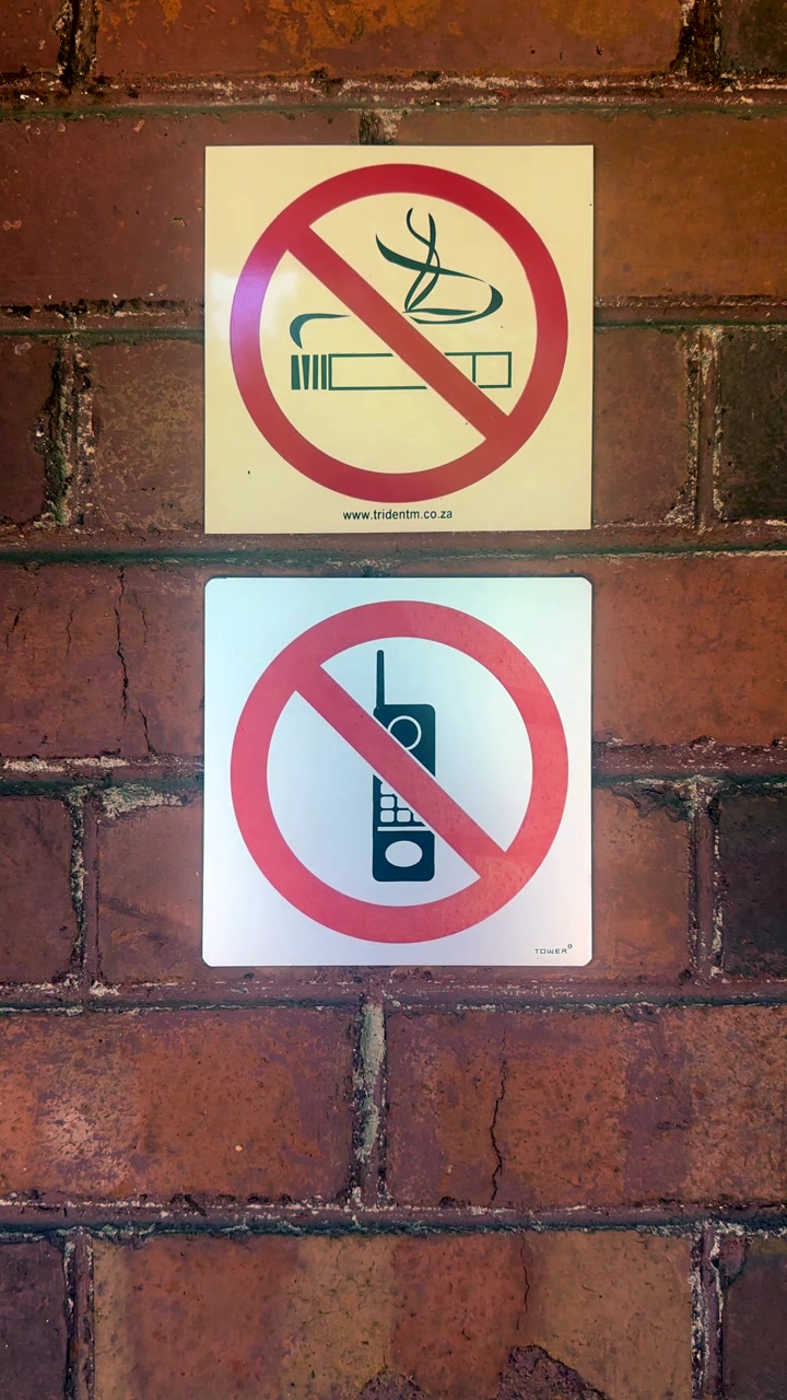 Signs prohibiting smoking and cell phone use are displayed on a brick wall. Useful for illustrating no-smoking areas or restricted zones in urban settings.
